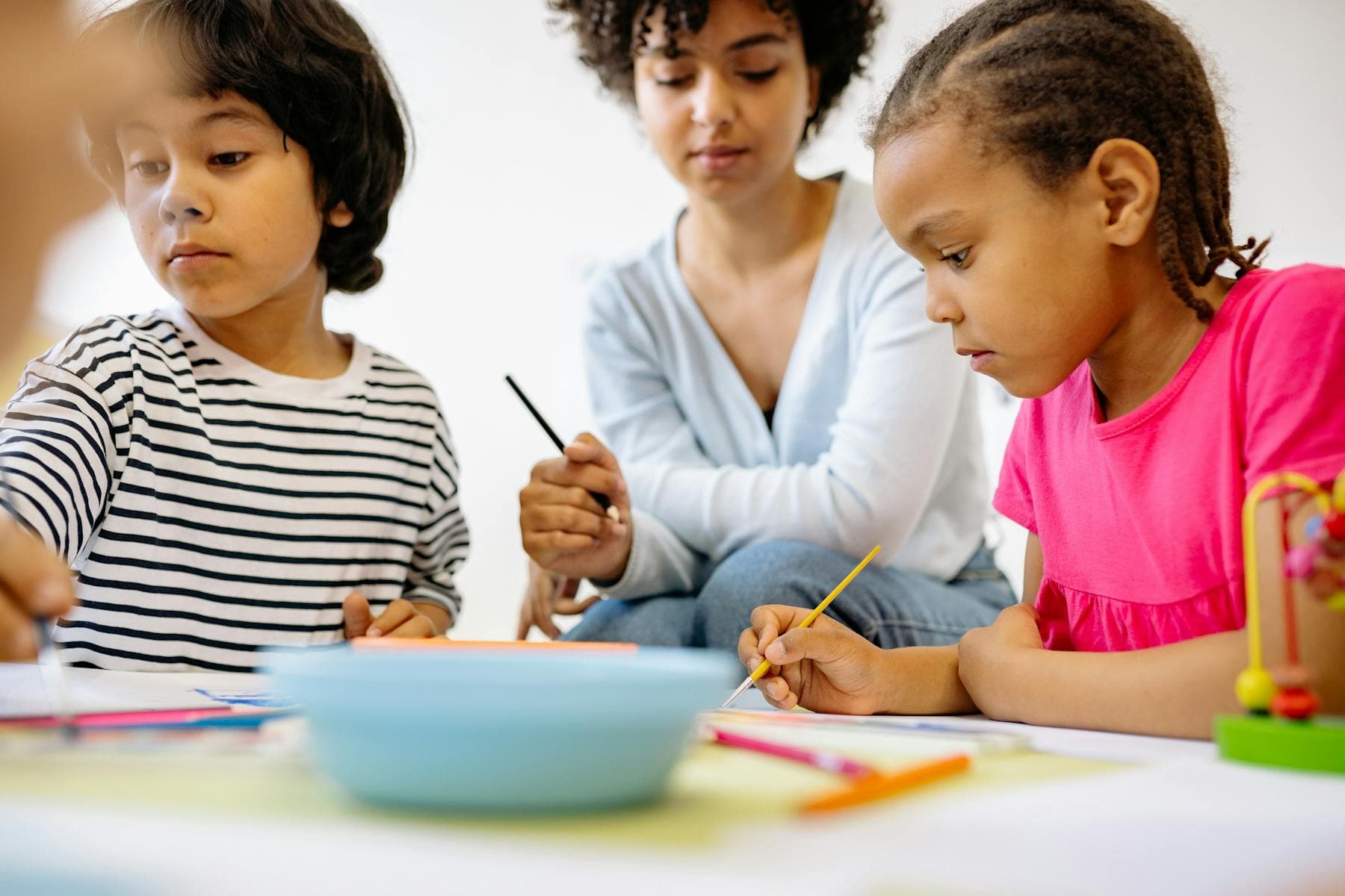 A teacher works with two young children at a painting and drawing table.