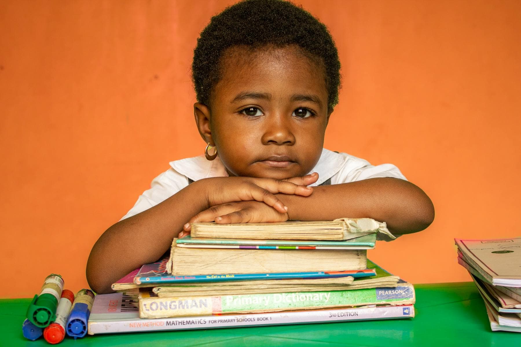 A young girl rests her chin on a stack of schoolbooks against an orange wall.